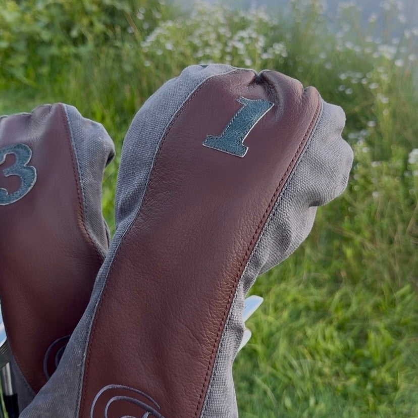 A close-up of golf club headcovers in brown and gray leather and fabric, with embroidered numbers and a monogram, photographed outdoors near a lake and grassy field.