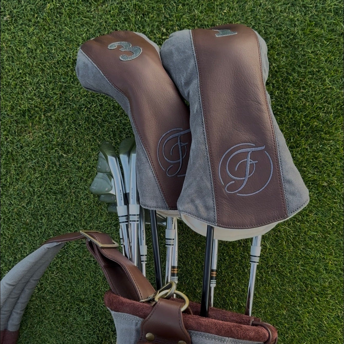 A golf bag filled with golf clubs, photographed on green grass. The bag and club heads are brown and gray with a white monogram logo.